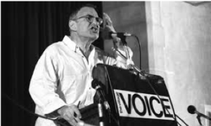 Larry Kramer at podium with The Village Voice Logo on the front