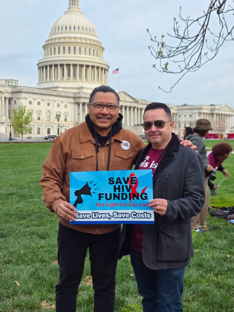Two men in front of the capitol building holding a sign saying "Save H.I.V. Funding"